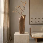Continuum hand-carved walnut wood sculpture vase displayed with a branch on a travertine pedestal in a quiet Japandi interior.