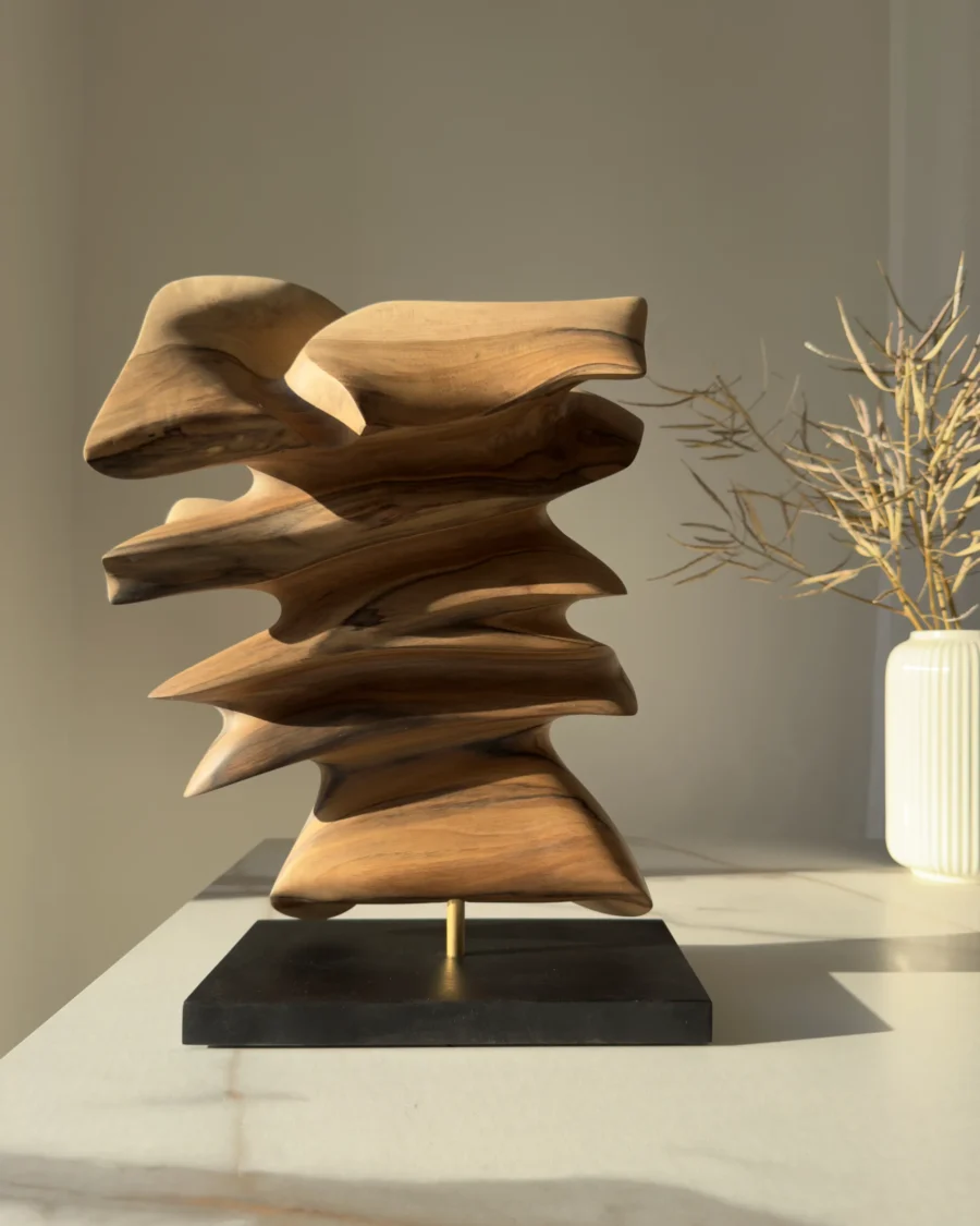 Fluens, a contemporary hand-carved walnut wood sculpture displayed in natural light beside a minimalist vase with branches.