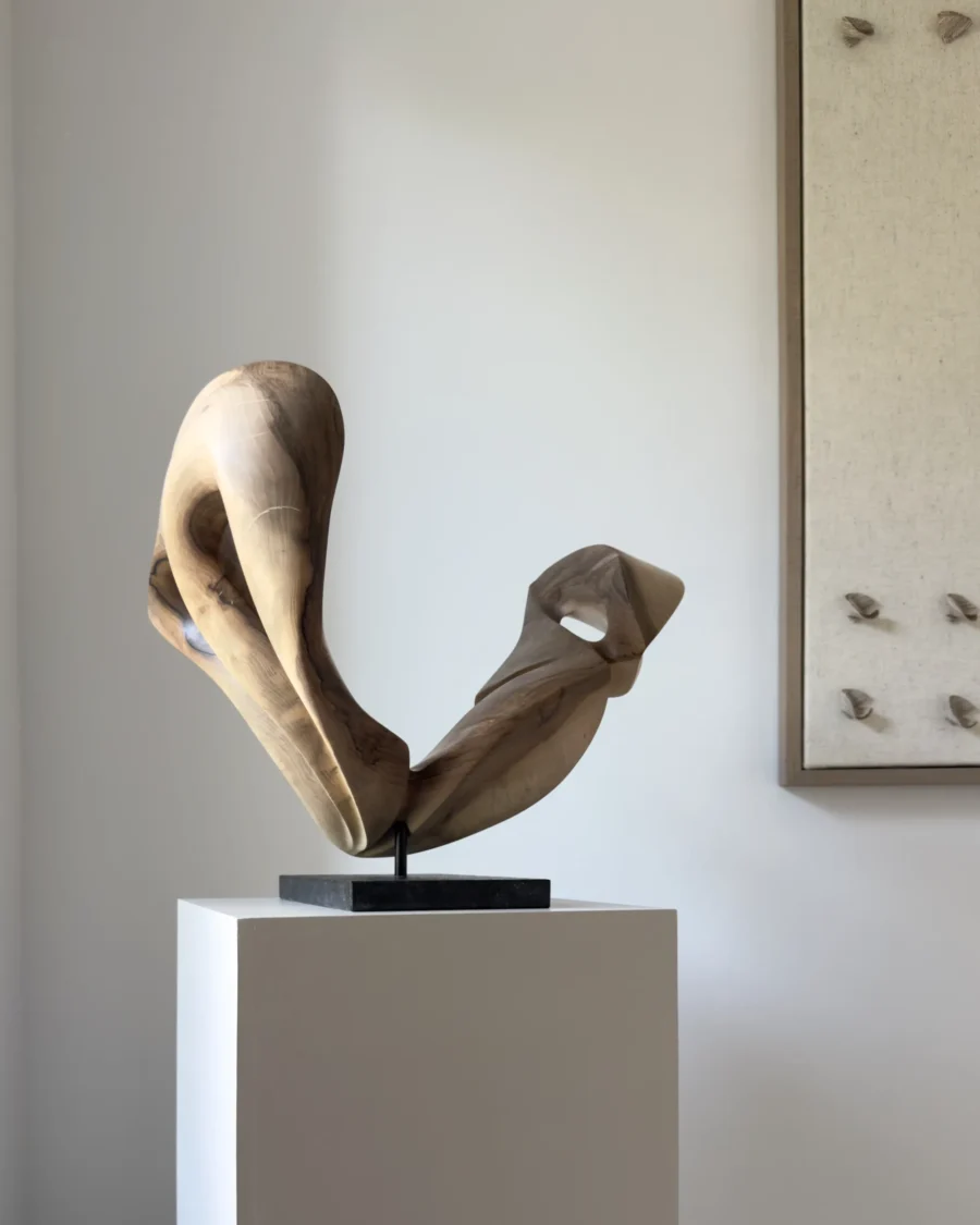 becoming-asymmetrical-walnut-sculpture-rear-view Becoming displayed on a pedestal beside a neutral textile artwork, showing how the asymmetrical walnut sculpture changes from a side angle.