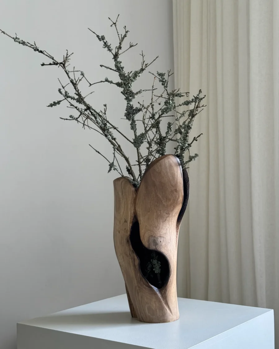Hollow hand-carved walnut vase holding delicate white dried flowers in a calm neutral interior.