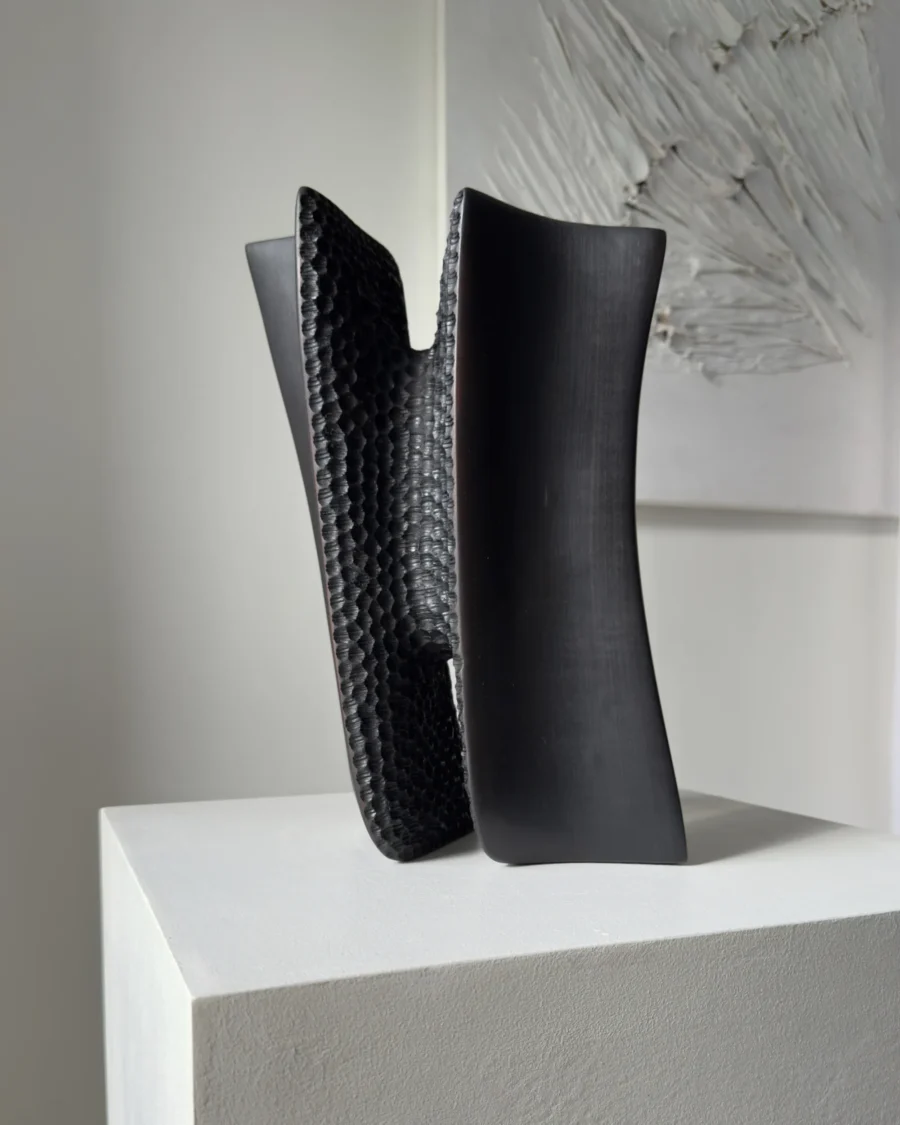 Pendere charred pear wood sculpture in its leaning position, showing smooth black surfaces contrasted with deep hand-carved texture.