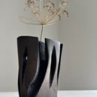 Construct sculptural wood vase holding a single dry stem, showing its dark architectural form and narrow central opening.