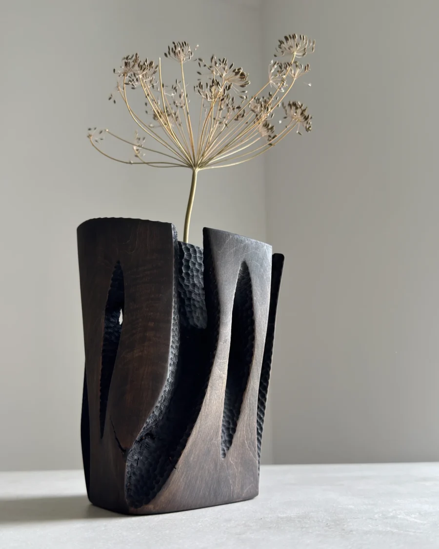 Construct sculptural wood vase holding a single dry stem, showing its dark architectural form and narrow central opening.