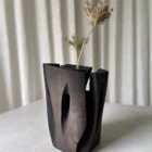 Construct dark wood vase with dry branches, showing its tall carved panels, open cracks and industrial-inspired structure.