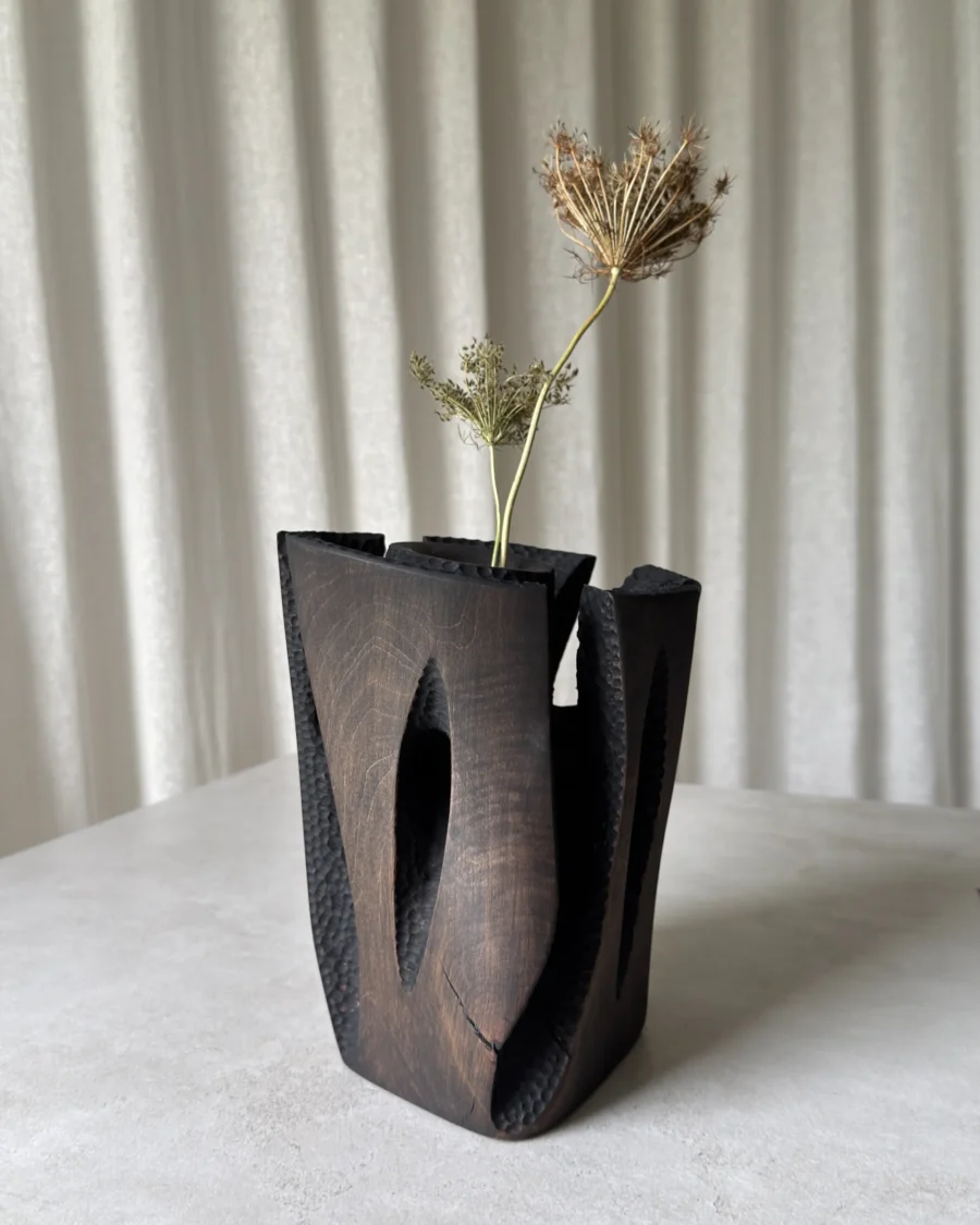 Construct dark wood vase with dry branches, showing its tall carved panels, open cracks and industrial-inspired structure.