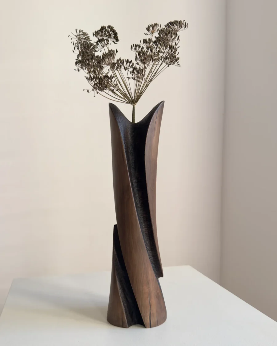 Tall sculptural cherry wood vase with delicate dried flowers, showing how botanical stems continue the upward movement of the carved form.